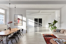 Tageslichtdurchflutetes Mietstudio in Berlin Treptow mit warmem Sonnenlicht, das durch große Fenster auf den weißen Boden und den Holztisch fällt. Im Raum: schwarze Stühle, Sofa mit rotem Teppich, Zimmerpflanze und dekorative Fotografie an der Wand.